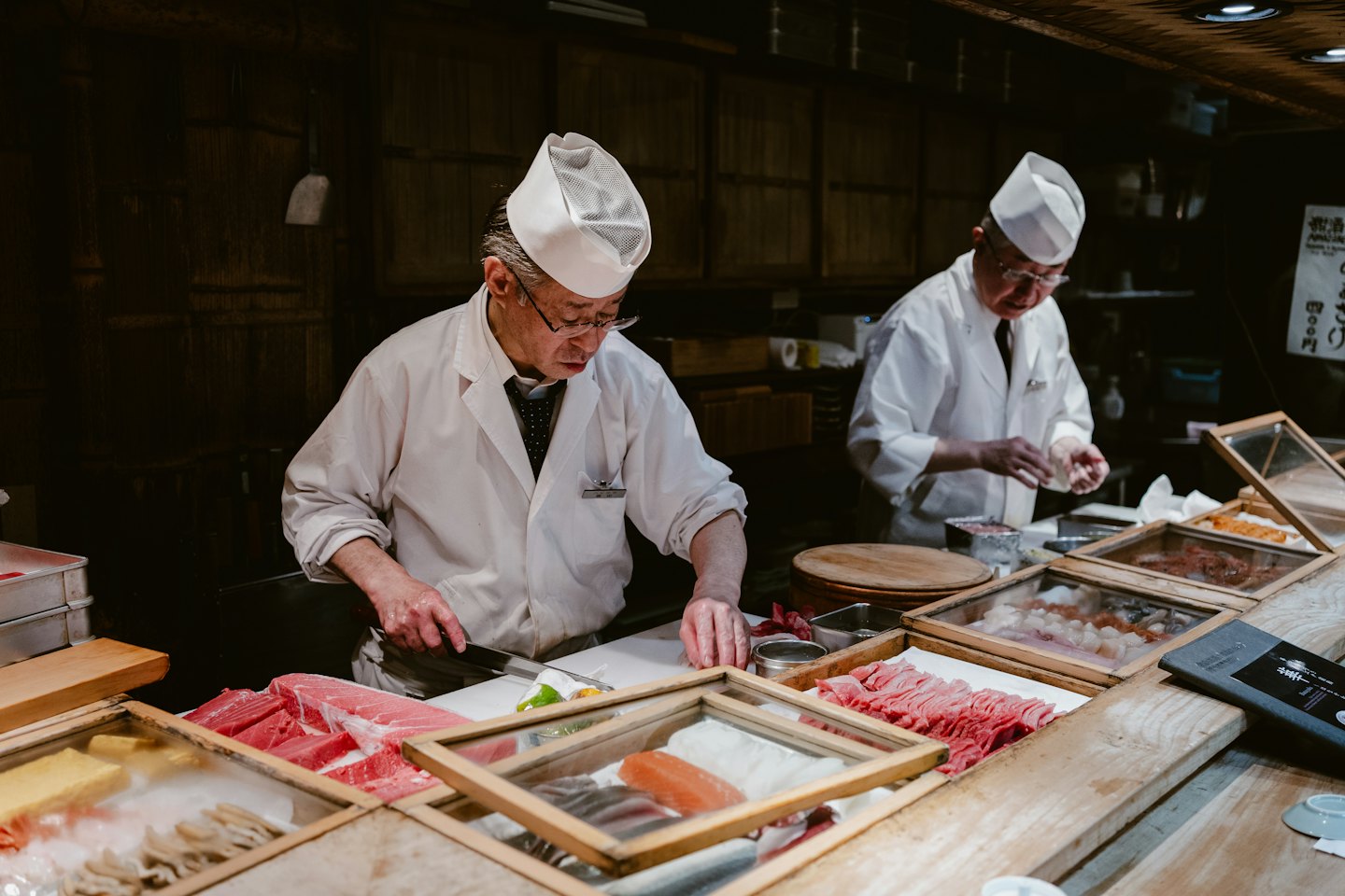 Two sushi chefs prepare raw fish behind a wooden counter.
