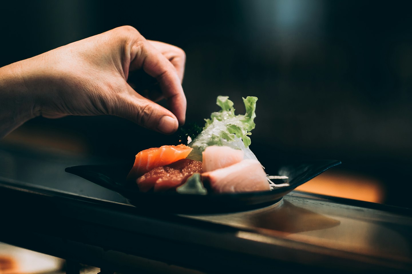 Chef's hand garnishing sashimi plate with salmon and wasabi.